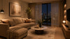 Cozy living room with a beige sofa, pillows, and a blanket, lit by a stylish lamp. Large window shows a city skyline at night. Warm, inviting ambiance.